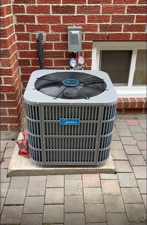 Air-conditioning condenser installed on a concrete pad beside a brick home, with service gauges connected.