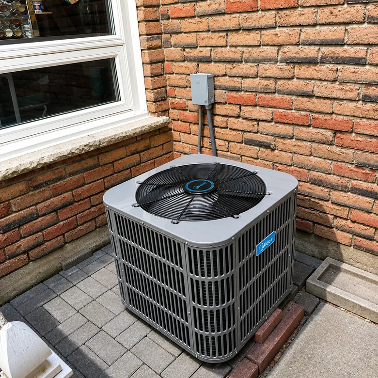 Newly installed Midea air-conditioning condenser on a paver pad beside a brick house, with the electrical disconnect on the wall above.